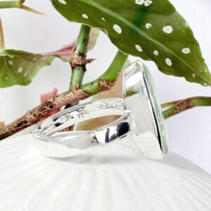May include: A sterling silver ring with a rectangular, light green gemstone. The ring is set against a backdrop of green leaves and white petals, highlighting the ring's polished silver band and the gemstone's clarity.