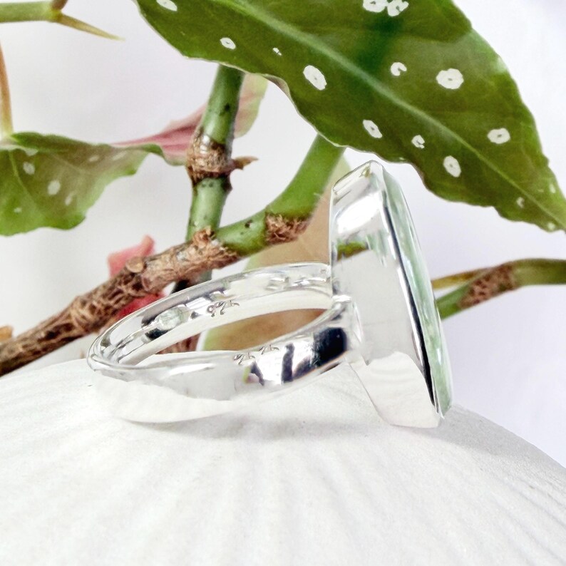 May include: A sterling silver ring with a rectangular, light green gemstone. The ring is set against a backdrop of green leaves and white petals, highlighting the ring's polished silver band and the gemstone's clarity.