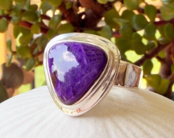 May include: A silver ring featuring a large, triangular, polished purple gemstone set in a smooth metal bezel. The stone displays deep violet hues with subtle, swirling patterns throughout its surface, held by a wide, solid silver band.