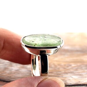 May include: A silver ring featuring a large, oval-shaped, pale green gemstone. The gemstone has a slightly translucent appearance with subtle variations in color. The ring's band and setting are polished silver, creating a contrast with the stone's color.