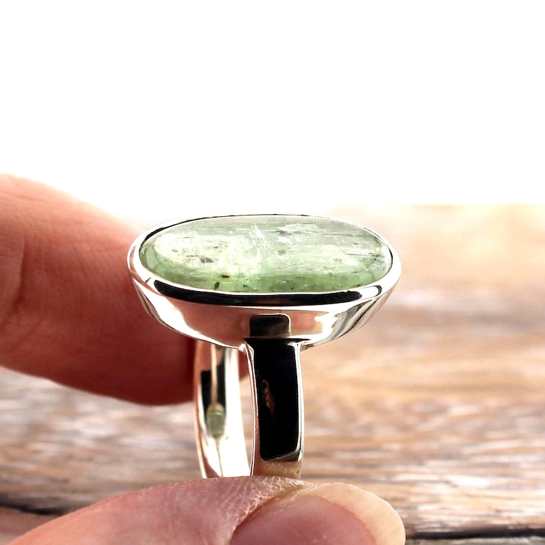 May include: A silver ring featuring a large, oval-shaped, pale green gemstone. The gemstone has a slightly translucent appearance with subtle variations in color. The ring's band and setting are polished silver, creating a contrast with the stone's color.