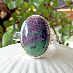 May include: A silver ring featuring a large, oval-shaped cabochon stone with a smooth, polished surface. The stone displays a distinct colour pattern with deep magenta-purple sections contrasting against vibrant green mineral inclusions throughout.