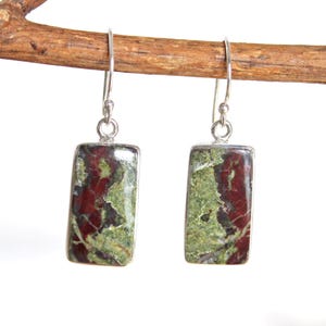 May include: A pair of rectangular earrings with a silver-colored metal frame. The earrings feature a natural stone with a mix of green and maroon colors. The earrings are hanging from a wooden branch.