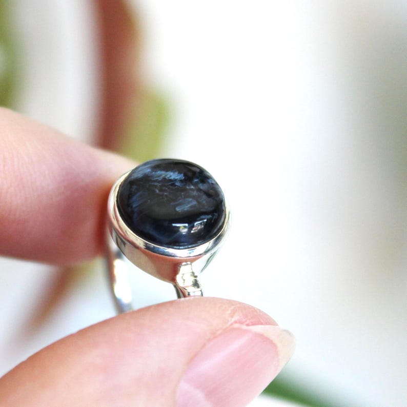 May include: A silver ring featuring a large, round, polished black gemstone with swirling patterns. The gemstone is set in a simple, elegant silver band. The ring is held up by a person's fingers.