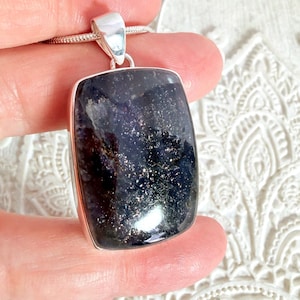 Peut inclure: Un pendentif rectangulaire avec une pierre bleu foncé mouchetée, sertie dans un cadre en métal argenté. Le pendentif est suspendu à une fine chaîne argentée. La pierre a un aspect scintillant et réfléchissant.