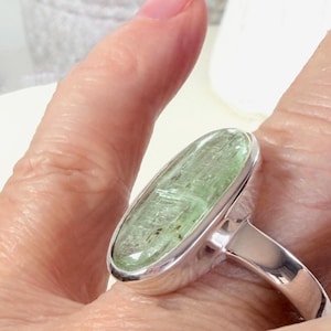 May include: A silver ring featuring a large, oval-shaped, light green gemstone. The gemstone has a textured appearance and is set in a polished silver band. The ring is shown on a finger.