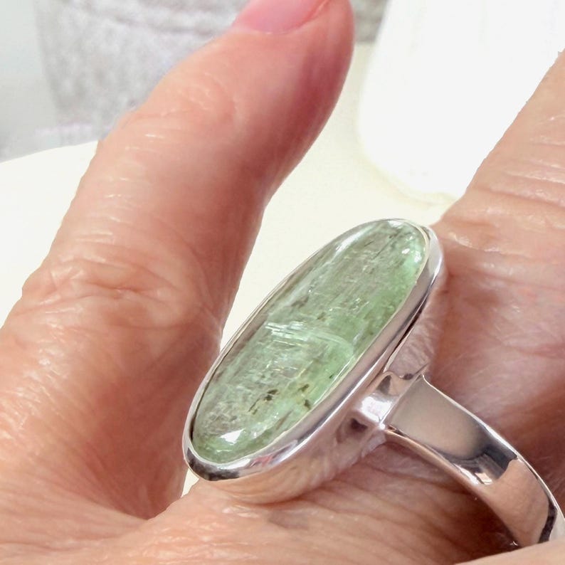 May include: A silver ring featuring a large, oval-shaped, light green gemstone. The gemstone has a textured appearance and is set in a polished silver band. The ring is shown on a finger.