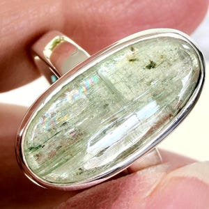 May include: A silver ring featuring a large, oval-shaped, pale green gemstone. The gemstone has a translucent quality with internal striations and subtle rainbow iridescence. The ring's band is a simple, polished silver design.