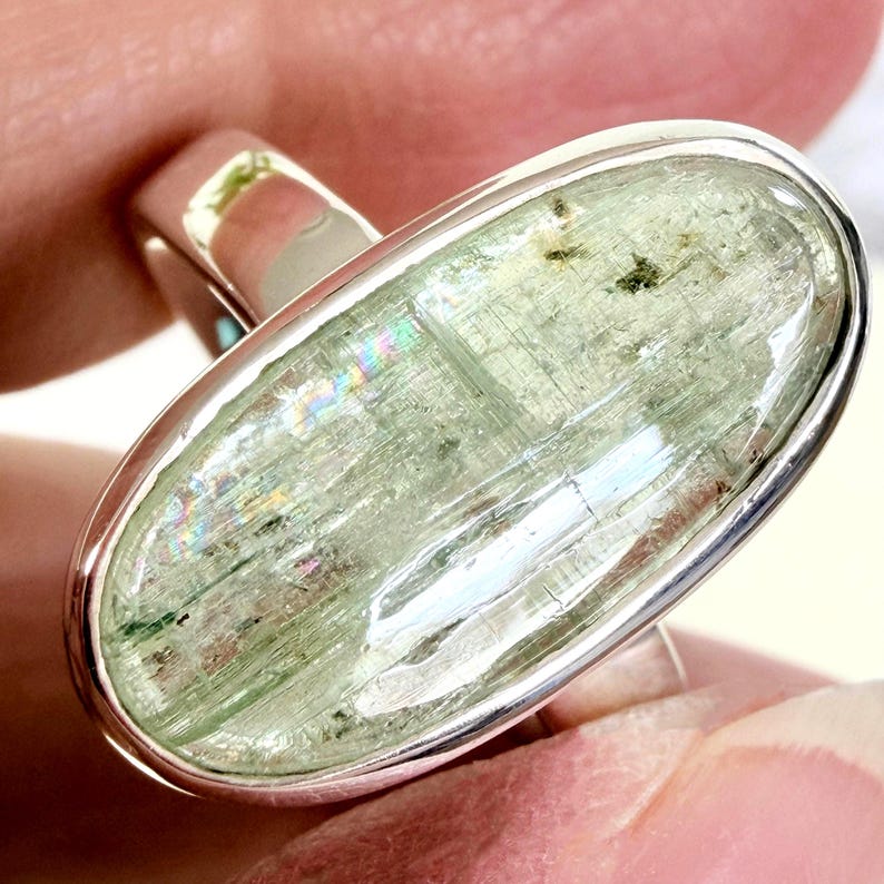 May include: A silver ring featuring a large, oval-shaped, pale green gemstone. The gemstone has a translucent quality with internal striations and subtle rainbow iridescence. The ring's band is a simple, polished silver design.