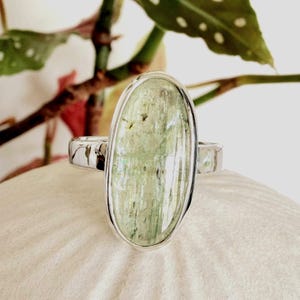 May include: A silver ring featuring a large, oval-shaped, pale green stone. The stone has a translucent appearance with subtle variations in color and texture. The ring's band is a polished silver, and the setting is a simple bezel design.