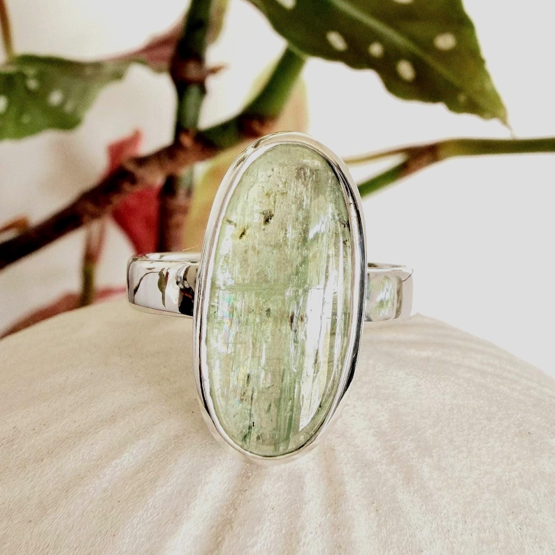 May include: A silver ring featuring a large, oval-shaped, pale green stone. The stone has a translucent appearance with subtle variations in color and texture. The ring's band is a polished silver, and the setting is a simple bezel design.