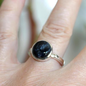 May include: A silver ring featuring a large, round, dark blue gemstone with intricate patterns. The gemstone is set in a simple, polished silver band. The ring is shown on a finger.