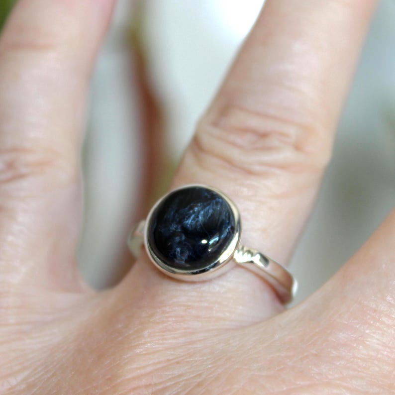 May include: A silver ring featuring a large, round, dark blue gemstone with intricate patterns. The gemstone is set in a simple, polished silver band. The ring is shown on a finger.