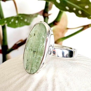 May include: A silver ring featuring a large, oval-shaped green gemstone with dark inclusions. The gemstone is set in a polished silver band. The ring is displayed against a white background with green foliage.