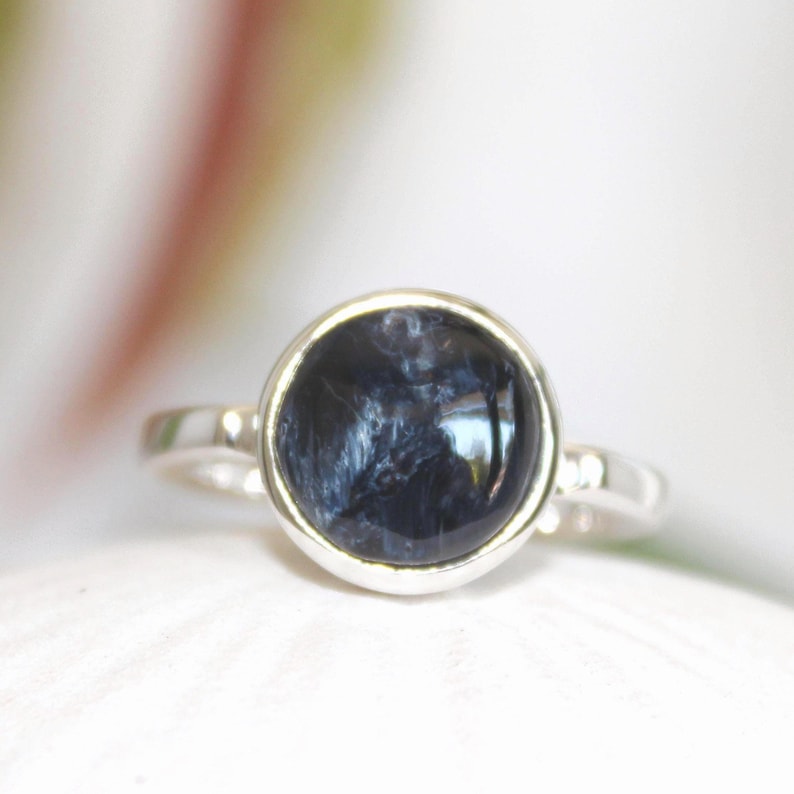 May include: A silver ring featuring a large, round, dark blue gemstone with swirling patterns. The gemstone is set in a bezel setting, and the ring has a simple, polished band. The ring is a piece of jewelry.
