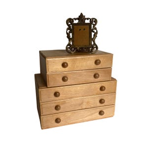 Peut inclure: Un petit meuble de rangement en bois clair avec cinq tiroirs et des boutons ronds en bois. Un cadre photo décoratif et orné de couleur bronze est posé dessus. Le cadre a un dos texturé.
