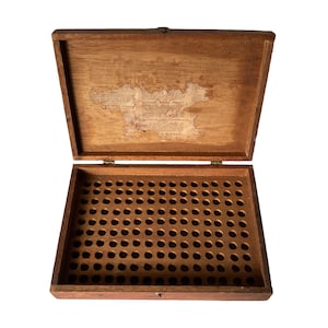 以下が含まれることがあります： ヒンジ付きの蓋が付いた開いた木製の箱。箱の内側には、小さな丸い穴のグリッドがあります。蓋には色あせた紙のラベルが付いています。箱は明るい茶色の木でできており、長方形の形をしています。