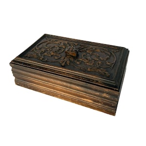 May include: A dark brown wooden box with an intricate carved floral design on the lid. The box has a raised, carved knob in the center of the lid.