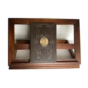May include: A dark brown wooden book stand with a rectangular frame and two small shelves. A vintage book with an embossed cover and a portrait medallion rests on the stand. The book's cover is dark brown with ornate floral designs.