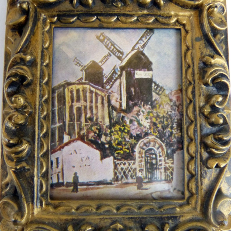 May include: A framed painting of a windmill in a rural setting. The windmill is painted in shades of brown and white, with a red roof. The painting is set against a backdrop of green trees and a blue sky. The frame is gold and ornate.