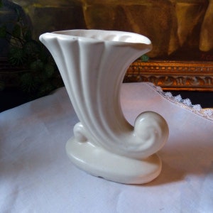 May include: A white ceramic vase shaped like a horn with a fluted design. The vase has a rounded base and a curled handle.