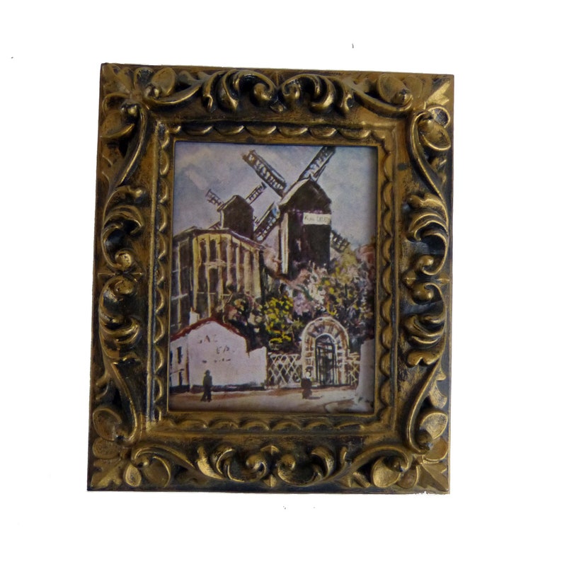 May include: A framed painting of a windmill in a European village. The painting is in a gold-colored frame with ornate details. The windmill is painted in shades of brown and gray, and the village is painted in shades of white, brown, and green.
