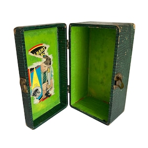 May include: An open, vintage green case with a textured exterior and a bright green interior. The inside of the lid features a torn advertisement with the text "ALBERT SC" and an illustration of a person using a microscope. The case has metal clasps.