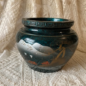 May include: A dark green ceramic planter with a gold and silver mountain scene and a black rim. The planter has a speckled finish.