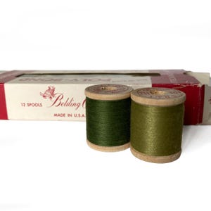 Könnte beinhalten: Zwei Spulen grünes Garn auf Holzspulen, neben einer Schachtel mit dem Text "Belding" und "Made in U.S.A.". Die Garnfarben sind Olivgrün und Waldgrün. Auf der Schachtel steht "12 Spulen".