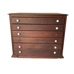 May include: A vintage wooden chest of drawers with six drawers. The chest is made of dark brown wood and features white, round knobs. The drawers are horizontally aligned and the chest has a rectangular shape.