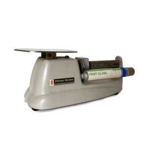 May include: A vintage light gray Pitney-Bowes postal scale with a flat metal weighing platform. The device features a horizontal cylindrical indicator with the text FIRST CLASS printed in green, and a metal scale bar with numerical markings.