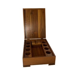 May include: Open wooden box with a hinged lid. The interior features multiple circular cutouts and a central rectangular compartment. The box is made of a dark brown wood with visible grain patterns, and is set against a white background.