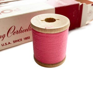 Könnte beinhalten: Eine Spule mit pinkfarbenem Faden und einem Holzkern, neben einer Vintage-Box. Auf der Box steht "Corticelli U.S.A. SINCE 1832". Der Faden ist eng um die Spule gewickelt.