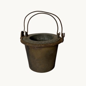 May include: A dark, weathered cast iron bucket with a tapered cylindrical body and a wide, flat rim. The bucket features a thin, arched metal wire handle attached to two side lugs. The surface shows signs of oxidation and a textured, matte finish.