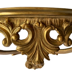 May include: A gold-painted, ornate shelf bracket with a decorative, scroll-like design. The shelf bracket has a curved top and a decorative base.