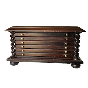 May include: A dark brown wooden coaster holder with six slots, each with a small brass knob. The sides feature a twisted column design, and the holder rests on four rounded feet. The top has a slightly overhanging edge.