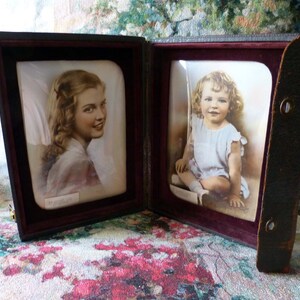 Può includere: Una cornice portafoto doppia vintage con due ritratti. La cornice è in pelle marrone scuro e ha una fodera in velluto bordeaux. Una foto è di una giovane donna, l'altra di un bambino piccolo. La cornice è aperta e in piedi.