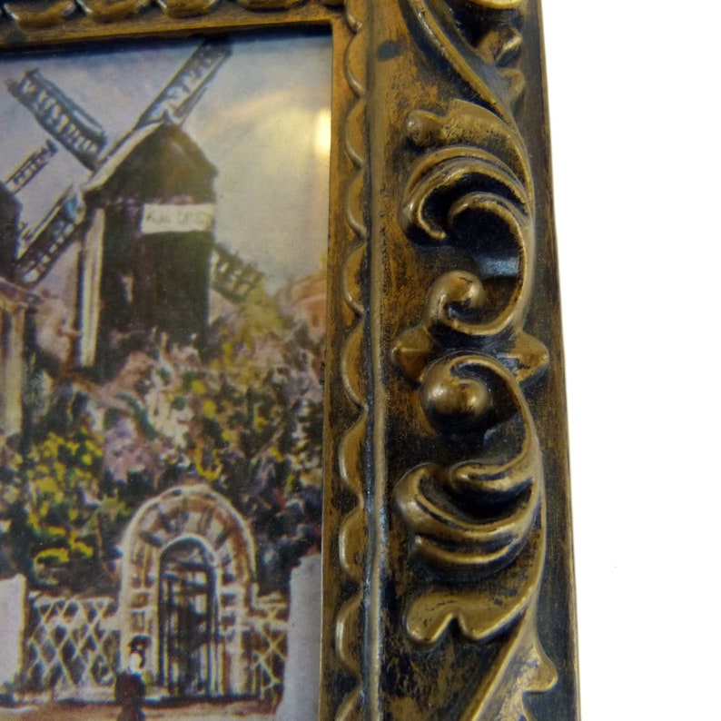 May include: A vintage-style picture frame with a gold-colored finish. The frame has a decorative, ornate design with a raised edge. The frame is empty and ready to display a picture or artwork.