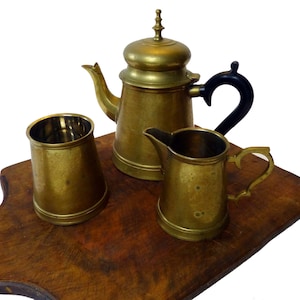 Può includere: Un set da tè in ottone con teiera, lattiera e zuccheriera. La teiera ha un manico nero e un pomello decorativo sul coperchio. Il set è esposto su un tagliere di legno.