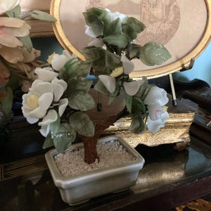 Pode incluir: Uma escultura de árvore bonsai em miniatura com flores brancas e folhas verdes. A árvore está em um vaso retangular de cerâmica verde claro preenchido com pequenas pedras brancas. O tronco é marrom.