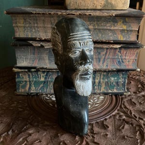 May include: A black stone bust sculpture of a man with a beard and a serious expression. The bust is sitting on a stack of old books.