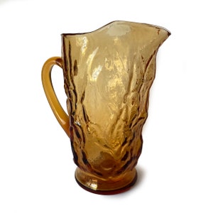Może przedstawiać: Dzbanek z bursztynowego szkła z fakturowanym wzorem przypominającym korę drzewa. Dzbanek ma szeroki otwór i uchwyt.