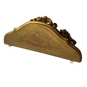 May include: A gold-painted wooden shelf bracket with ornate carvings and a curved top. The bracket has two metal rings on the bottom for hanging.