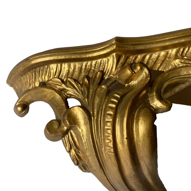 May include: A gold-painted, ornate wall shelf with a decorative, scrolled design. The shelf is made of wood and has a smooth, polished finish.