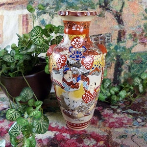 May include: A decorative, hand-painted ceramic vase with a detailed design featuring figures in traditional clothing. The vase has a cream-colored base with red, yellow, blue, and brown accents. The vase is approximately 10 inches tall.