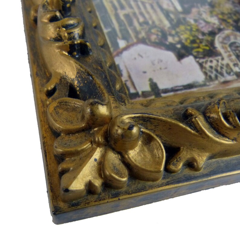 May include: Close-up of a gold-colored ornate picture frame with a black distressed finish. The frame has a detailed floral design.