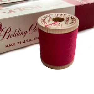 Könnte beinhalten: Eine Holzspule mit leuchtend rotem Faden, auf der "Belding Corticelli" und "Poly-Bond" aufgedruckt sind. Daneben eine Schachtel mit den Aufdrucken "Belding Co" und "Made in U.S.A. Since". Der Faden ist sauber um die Spule gewickelt.