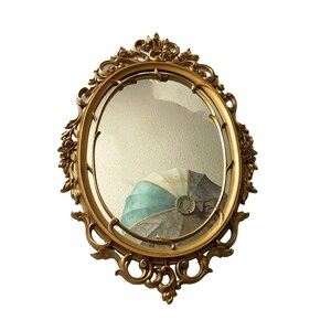 May include: A gold oval mirror with an ornate, carved frame. The mirror reflects a blue and white patterned object.