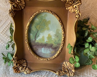 May include: A gold-toned ornate frame with a painting of a landscape with a waterfall. The frame has a decorative design with scrolls and swirls.
