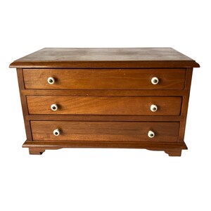May include: A small, three-drawer wooden chest with a rich, warm brown finish. Each drawer features a white, round knob. The chest has a simple, classic design, suitable for storing small items.